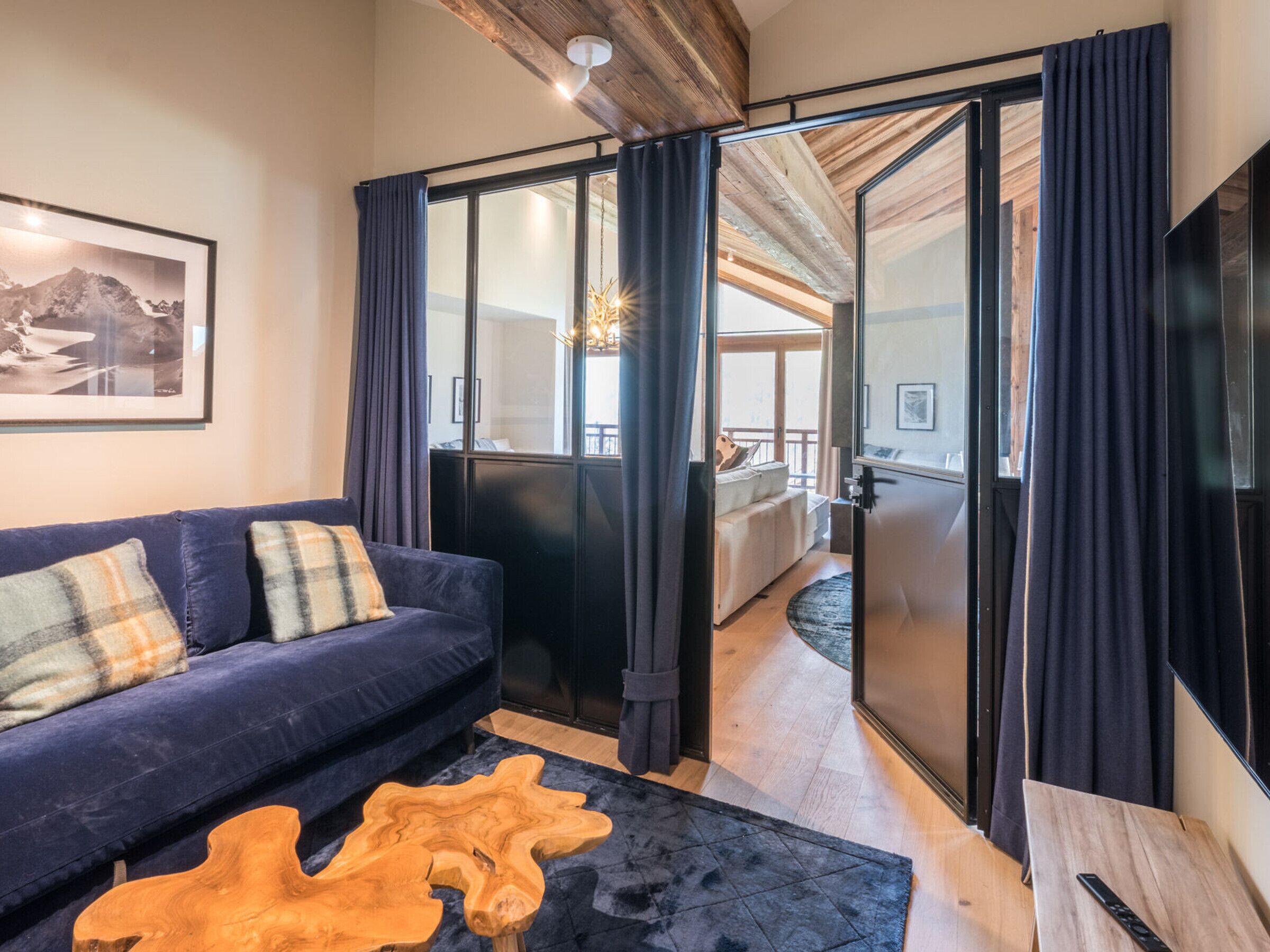Una sala de estar moderna con un sofá azul, una mesa de centro de madera y vista a un dormitorio a través de una puerta de vidrio.