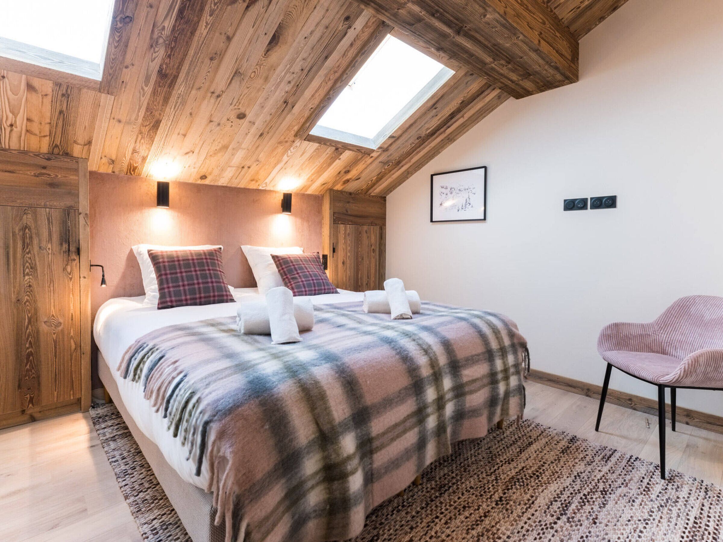 A cozy bedroom with a wooden ceiling, a large bed with plaid bedding, a chair, and a skylight.