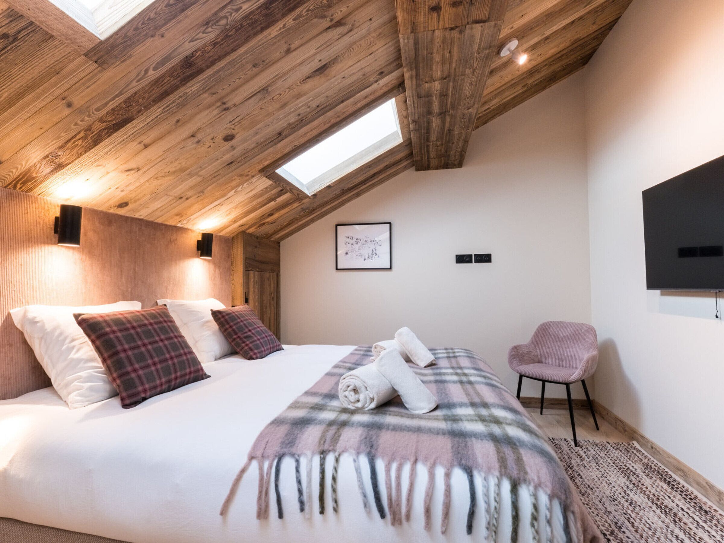 A bedroom with a wooden ceiling, a bed with white and plaid bedding, a chair, a flat-screen TV, and a framed picture on the wall.