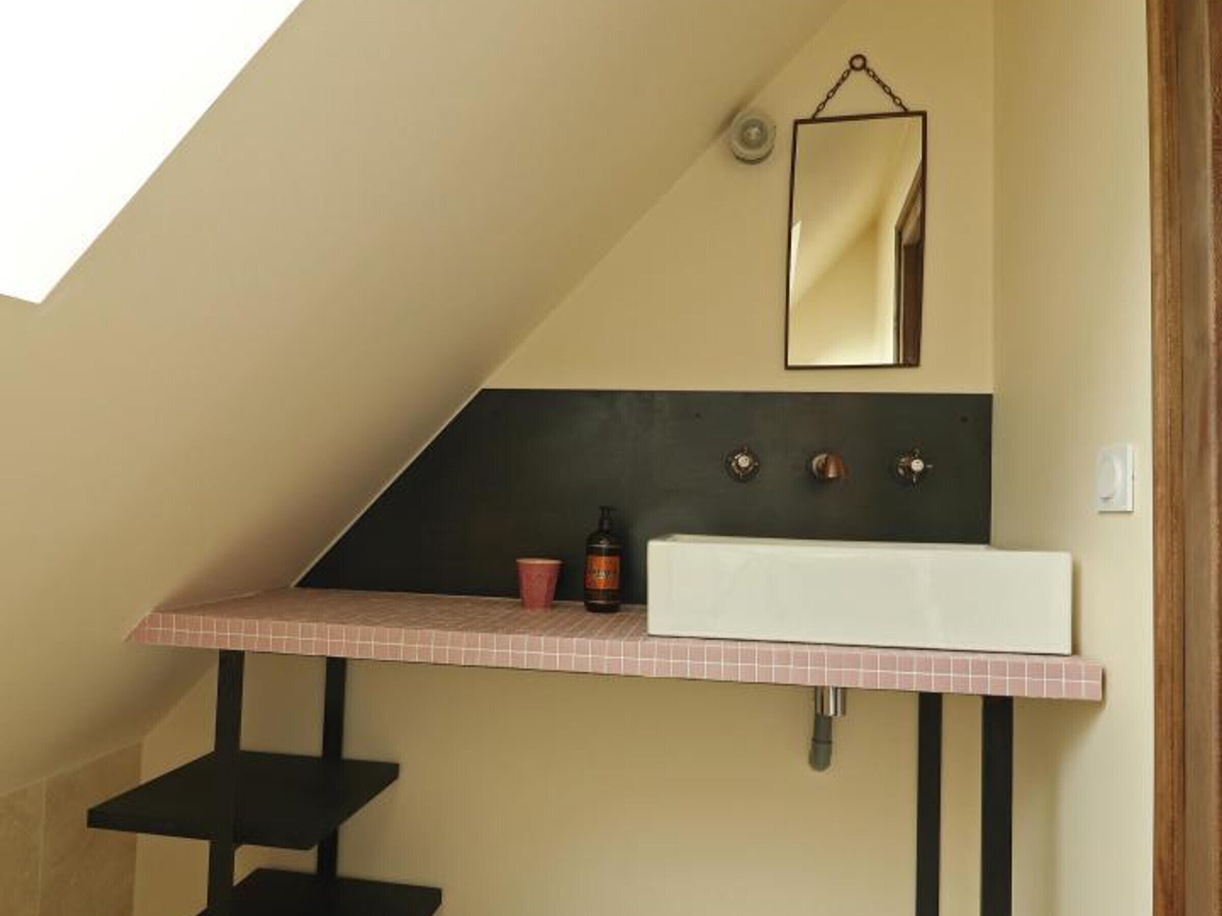 Une salle de bain avec un plafond en pente, un lavabo blanc, un miroir et une bouteille de shampoing.