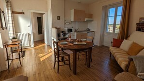 Een gezellige woonkamer met een houten vloer, een eettafel met stoelen, een bank en een keuken met kastjes en een raam.