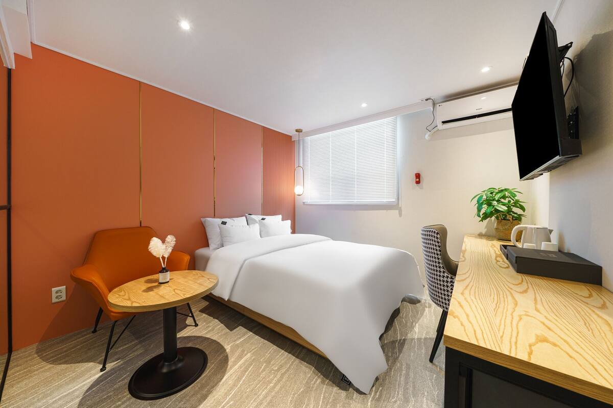 A modern hotel room with a bed, orange armchair, round wooden table, and a TV mounted on the wall.