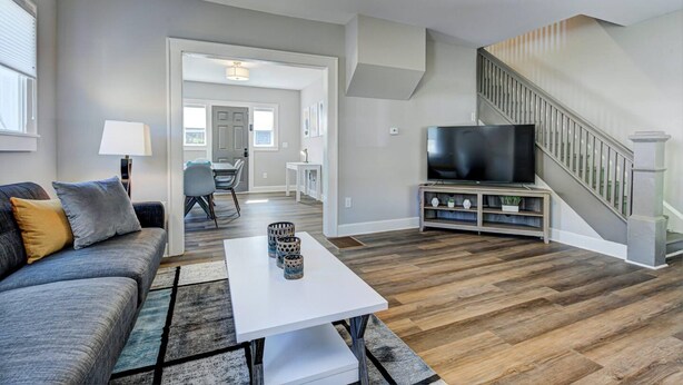 A modern living room with a grey sofa, a white coffee table, and a TV on a wooden stand. A staircase is visible in the background.