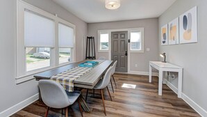 A dining area with a long table, chairs, and a bench. There are windows with blinds, a door, and a wall-mounted light fixture.