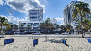 Een strand met blauwe ligbedden, palmbomen en moderne gebouwen op de achtergrond.