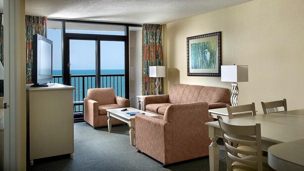 A hotel room with a balcony, a sofa, a dining table, and a TV. - Beaming Oceanfront Three Bedroom Condo: 7th Floor (Myrtle Beach)