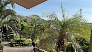 A balcony with a patterned cushion, metal railing, and a view of a garden with palm trees and a building in the background. - Happy Oasis Second Floor Condo with Terrace (Playas Del Coco)