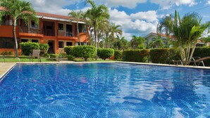 A swimming pool with clear blue water, surrounded by palm trees and a building with balconies. - Happy Oasis Second Floor Condo with Terrace (Playas Del Coco)