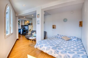 Quarto moderno com uma cama grande, uma mesinha e um sofá. Cozinha visível ao fundo.