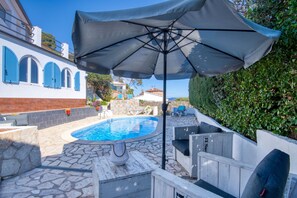 A swimming pool with a stone deck, surrounded by outdoor furniture and a large umbrella.