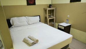 A neatly made bed with white linens and two rolled towels, a bedside table with a water bottle and a phone, and a small nightstand with a vase of flowers. - Promosia Guest House (Surabaya)