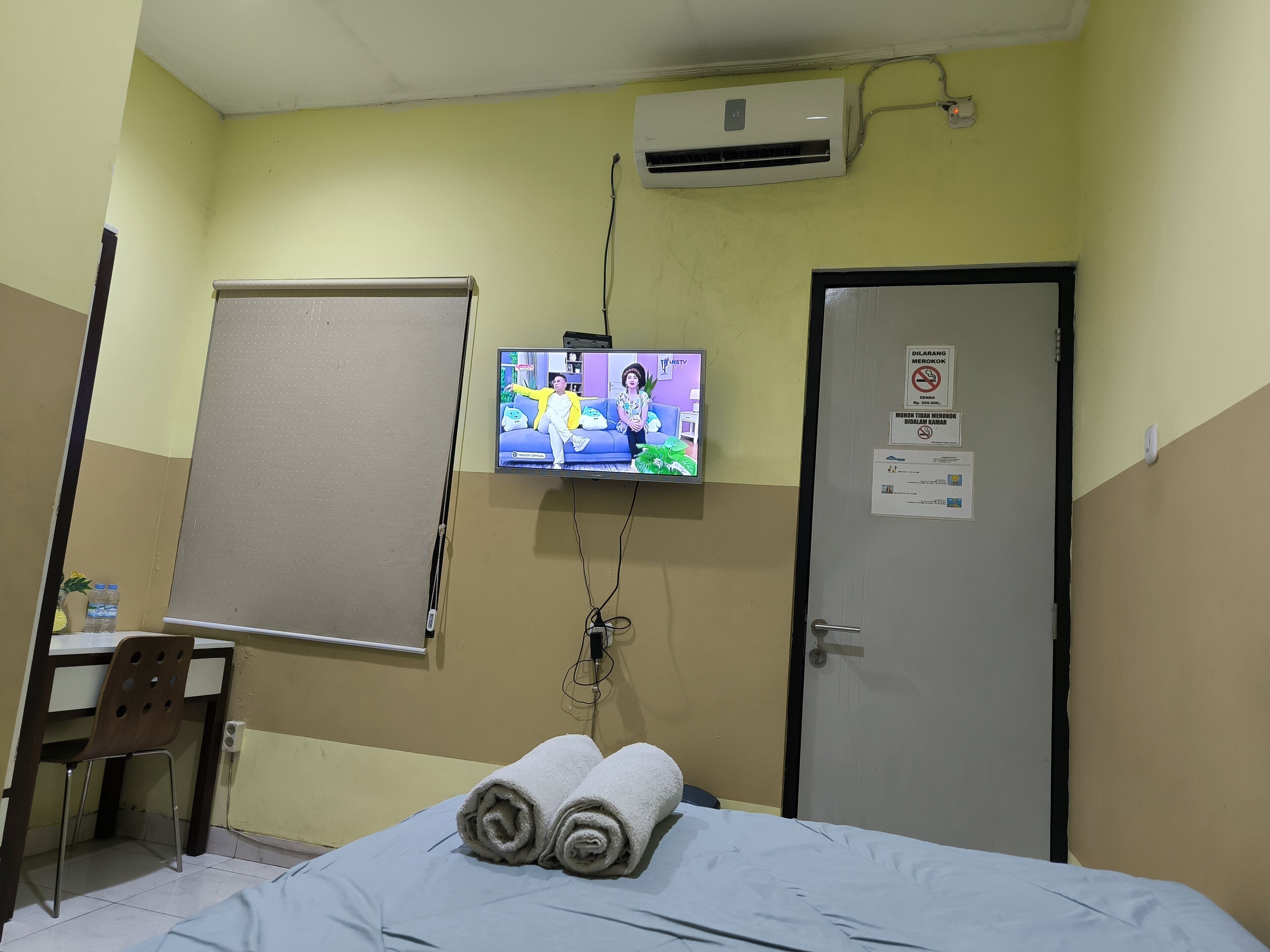 A hotel room with a bed, a desk, a chair, an air conditioner, a TV, and a door with a sign.
