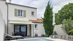 A swimming pool with a wooden deck, a covered patio with outdoor furniture, and a tall evergreen tree. - Maison de Flora à 8mn des Plages (Arces)