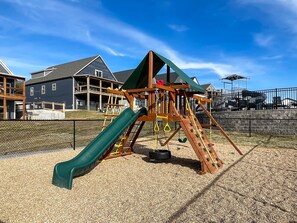 A playground with a slide, climbing net, and swings, surrounded by a fence and houses in the background.