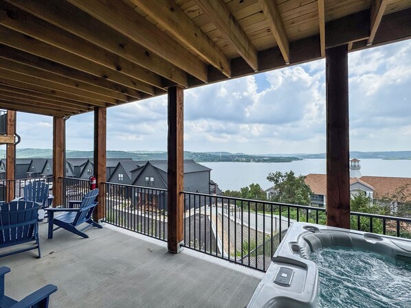 A covered balcony with Adirondack chairs, a hot tub, and a view of a lake and buildings.