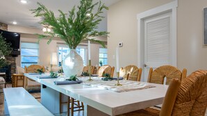 A dining area with a long table, wicker chairs, and a large vase with greenery.