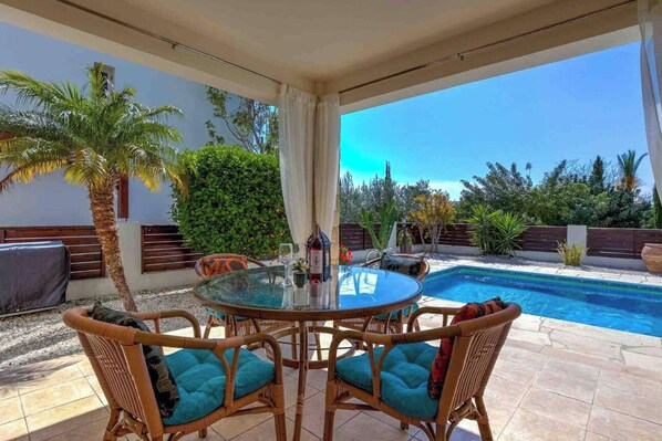 Eine Terrasse im Freien mit einem runden Tisch, Korbstühlen, einem Pool und umgebender Bepflanzung.