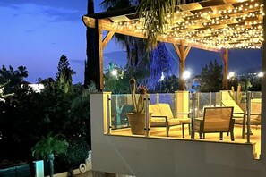 Een modern terras met een glazen balustrade, comfortabele zitplaatsen en een plafond versierd met lichtslingers.