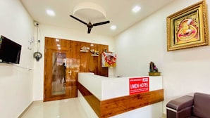 A hotel reception area with a wooden reception desk, a ceiling fan, a television mounted on the wall, and a framed artwork above the desk. - Hotel Vistacrest Noida Sector 104 (Noida)