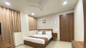 A bedroom with a bed, curtains, a ceiling fan, a bedside table, a nightstand, a wall-mounted light, and a door. - Hotel Vistacrest Noida Sector 35 (Noida)