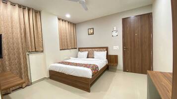 A bedroom with a bed, curtains, a ceiling fan, a bedside table, a nightstand, a wall-mounted light, and a door.