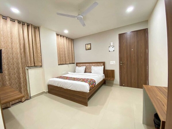 A bedroom with a bed, curtains, a ceiling fan, a bedside table, a nightstand, a wall-mounted light, and a door. - Hotel Vistacrest Noida Sector 35 (Noida)