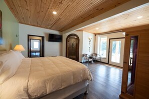 A bedroom with a large bed, wooden ceiling, a TV mounted on the wall, and a door leading to another room.