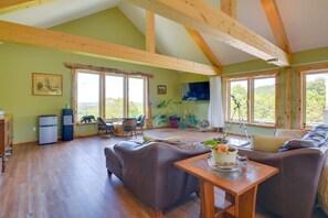 A spacious living area with a high ceiling, wooden beams, a sofa, a table with a cake, and large windows offering a view of the outdoors. - Private Pool & Hot Tub | Fall color view for miles (Eureka Springs)