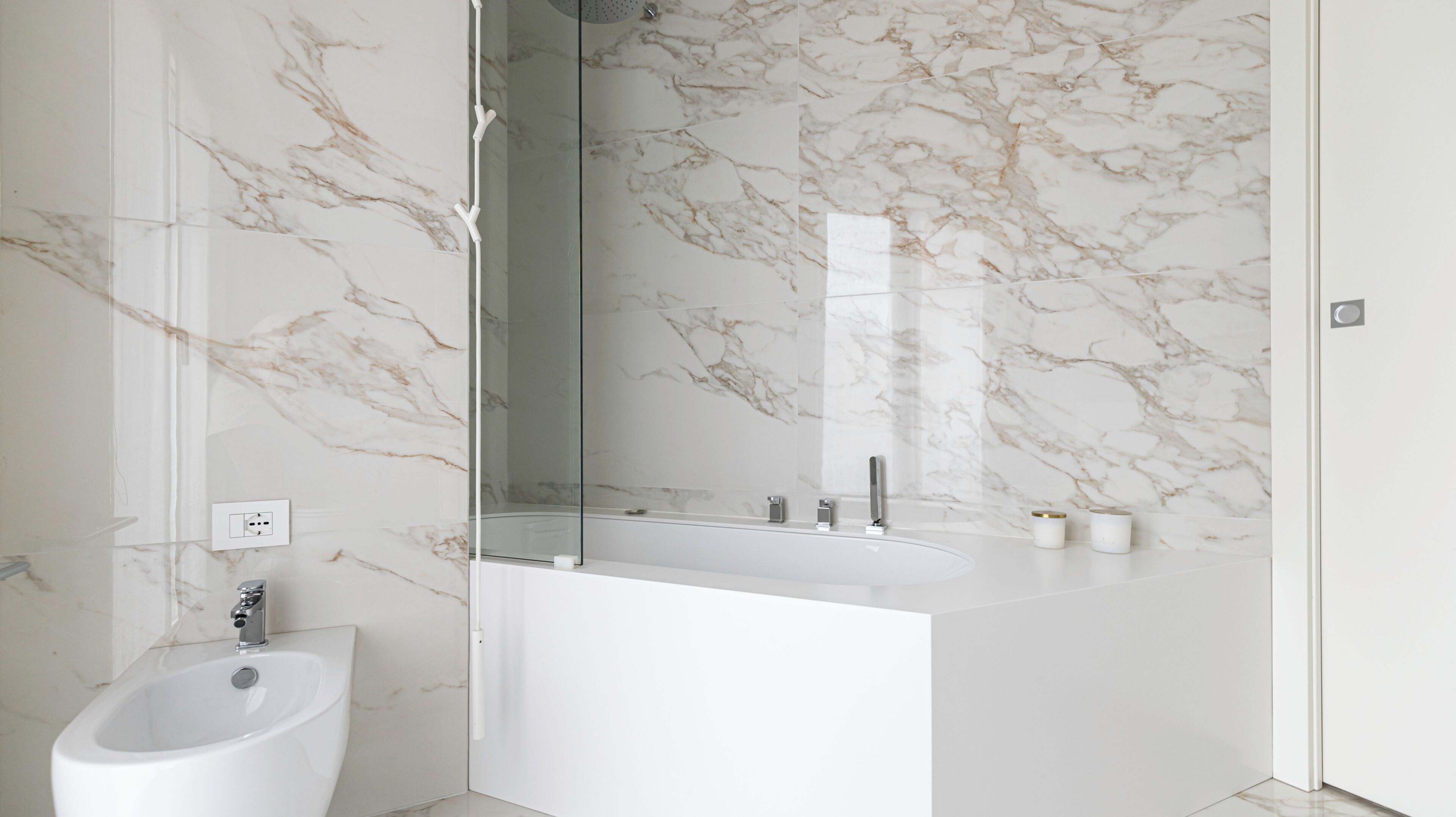 Une salle de bain moderne avec un mur en marbre, une douche à parois de verre, un lavabo blanc et un bidet.