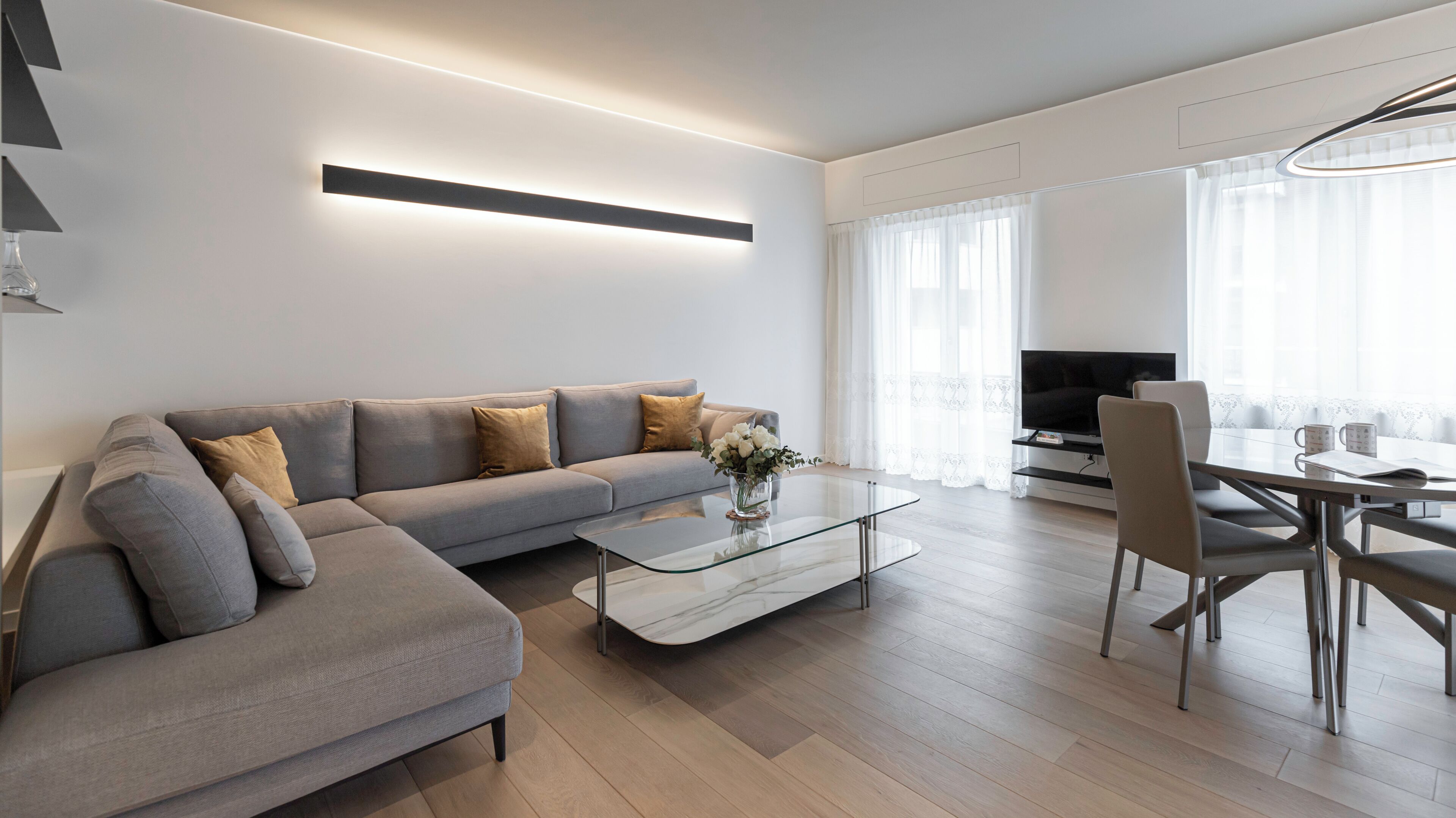 A modern living room with a grey sofa, a glass coffee table, and a dining area with a TV.