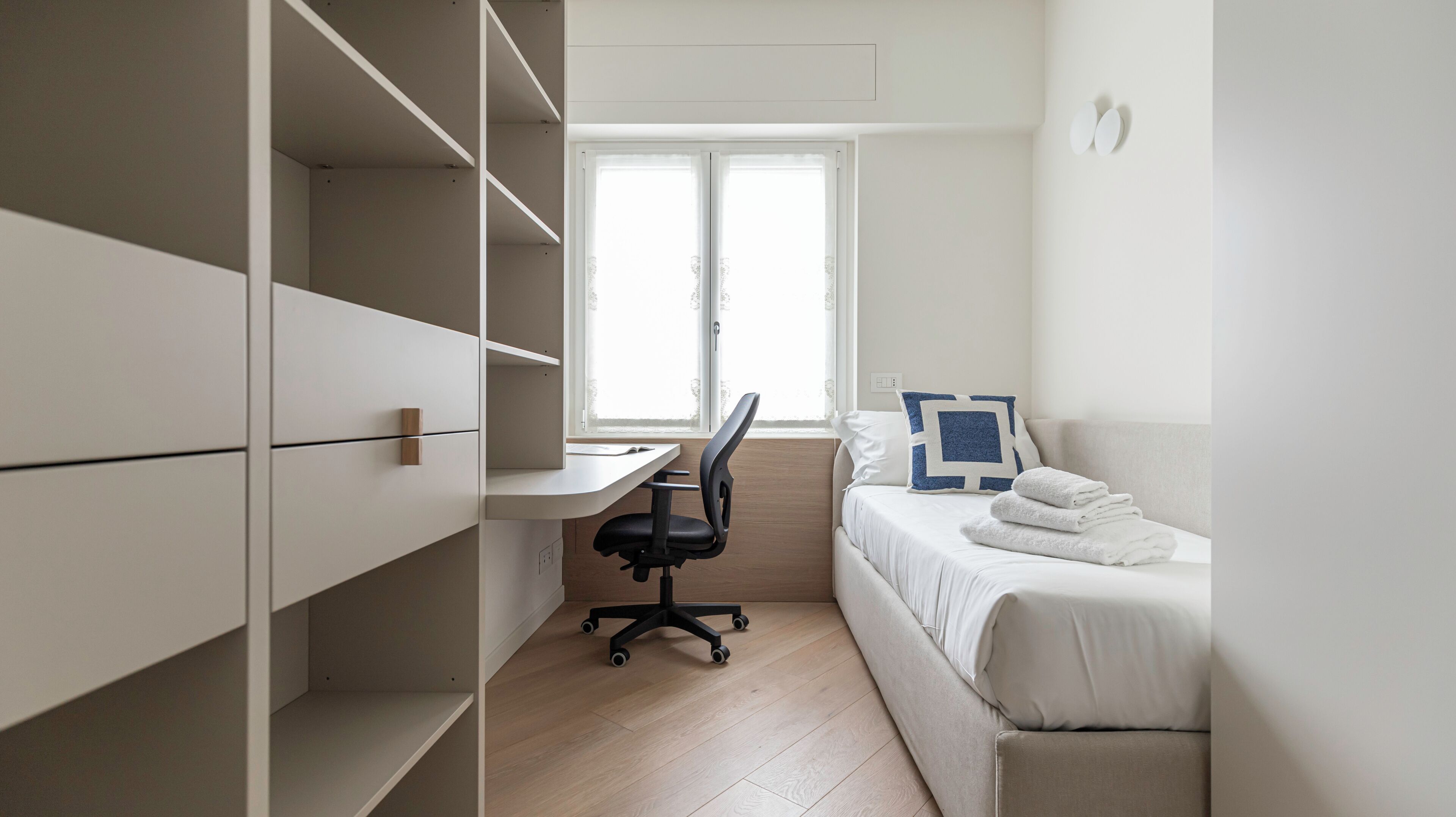 A modern, minimalist room with a desk, chair, and a bed with neatly folded towels.