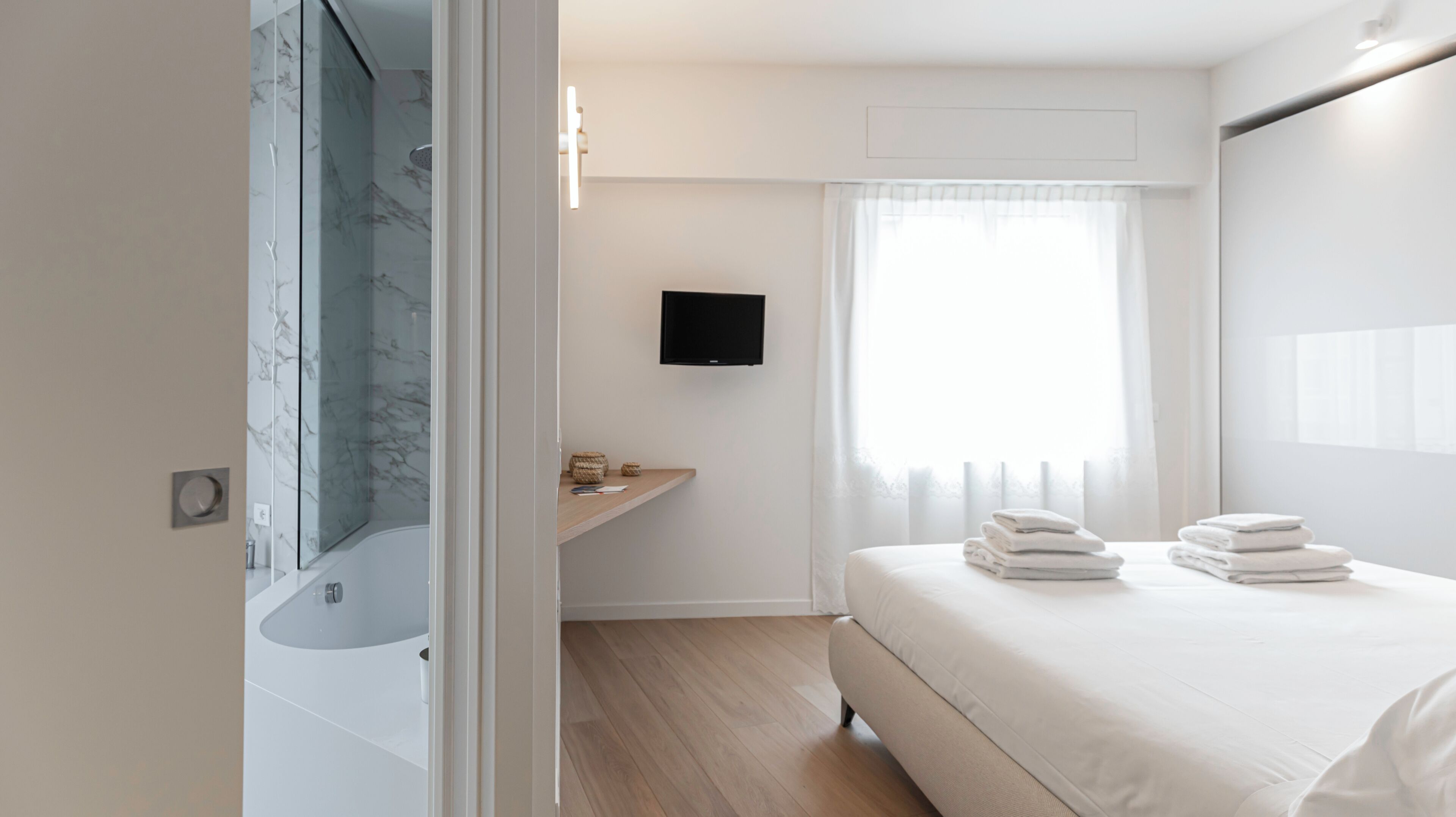 A modern hotel room with a marble shower, a flat-screen TV, a wooden vanity, and a neatly made bed with folded towels.