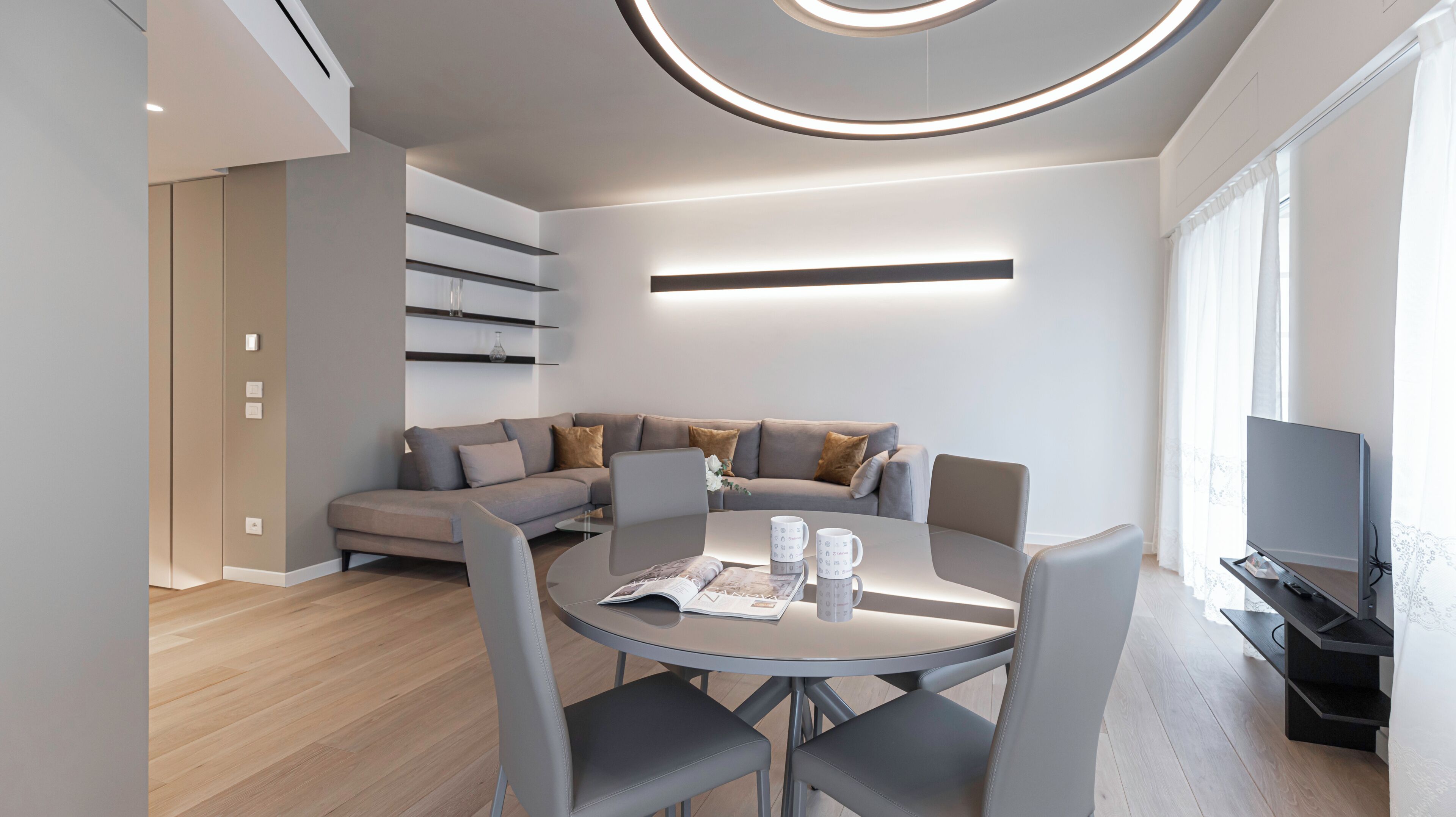 A modern living room with a grey sofa, a round dining table, and a flat-screen TV.
