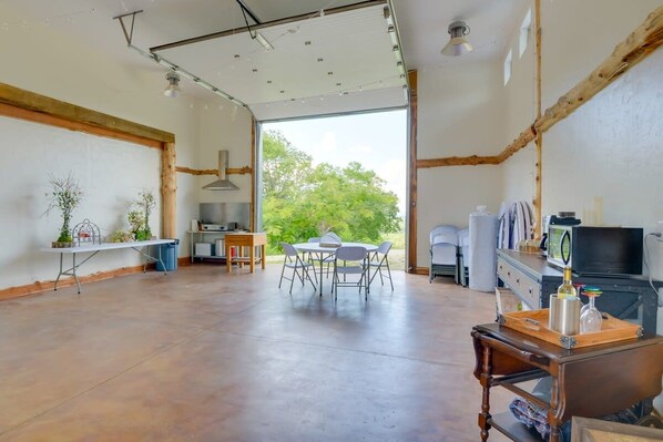 A spacious room with a large table and chairs, a kitchen area, and a view of greenery outside.