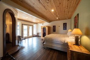 Une chambre spacieuse avec un grand lit, un plafond en bois et une vue sur l’extérieur, visible à travers une porte en arc de cercle.