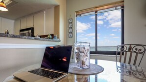 A laptop on a glass table with a clear vase of seashells, a view of the sky through a window, and a kitchen in the background. - Million Dollar Views! 1B/2B Bayfront Windward Point 508 (Fort Myers Beach)