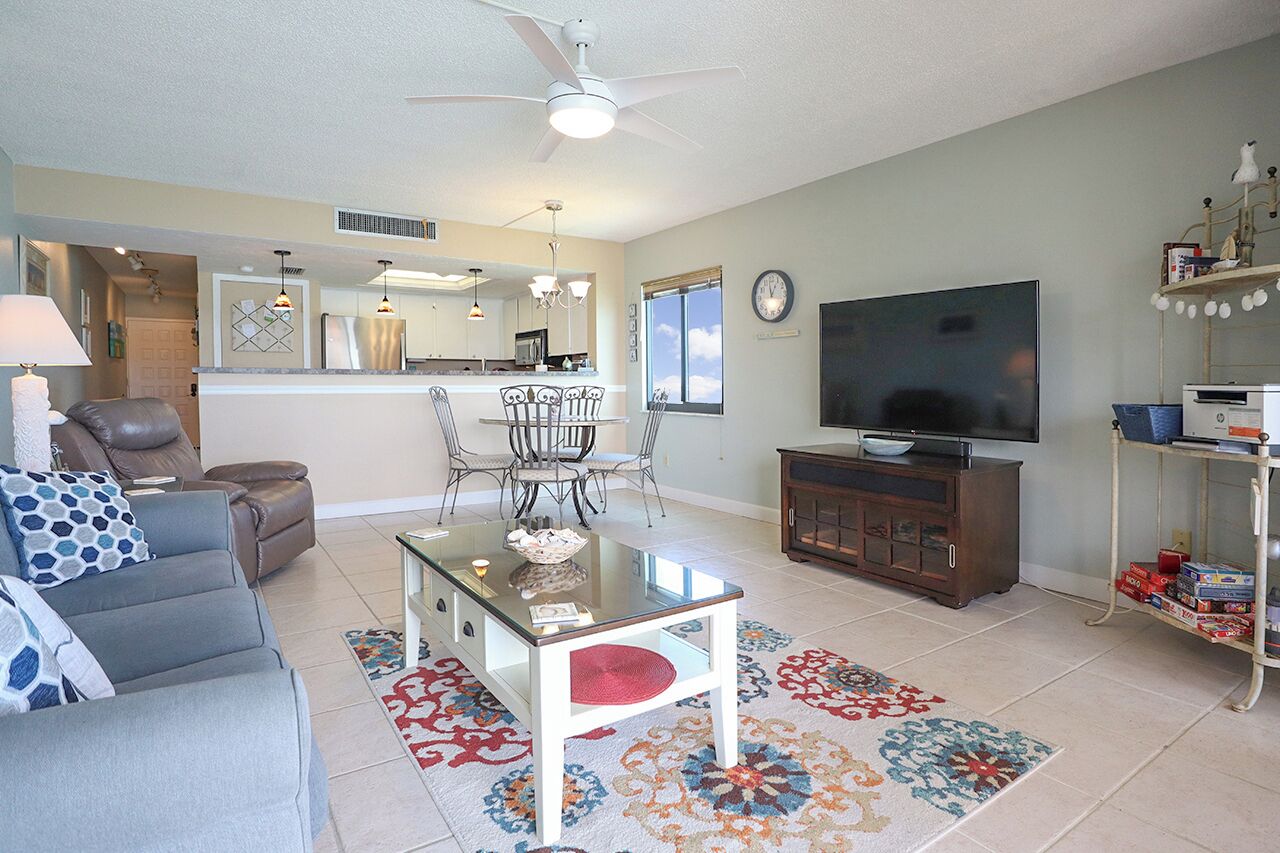 A living room with a sofa, a glass coffee table, a TV, a dining area with chairs, a kitchen in the background, and a ceiling fan.