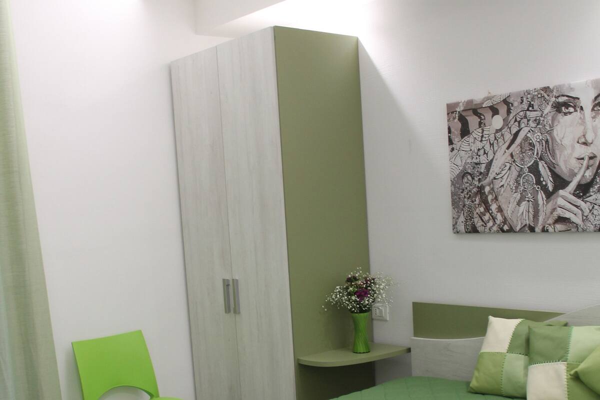 A neatly made bed with a green bedspread, a green chair, a wardrobe, and a painting on the wall.