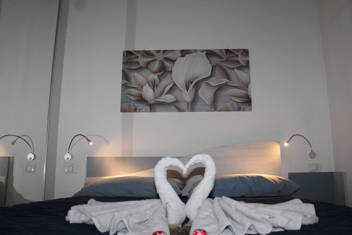 A hotel room with a bed, a heart-shaped towel arrangement, and a floral wall art piece.