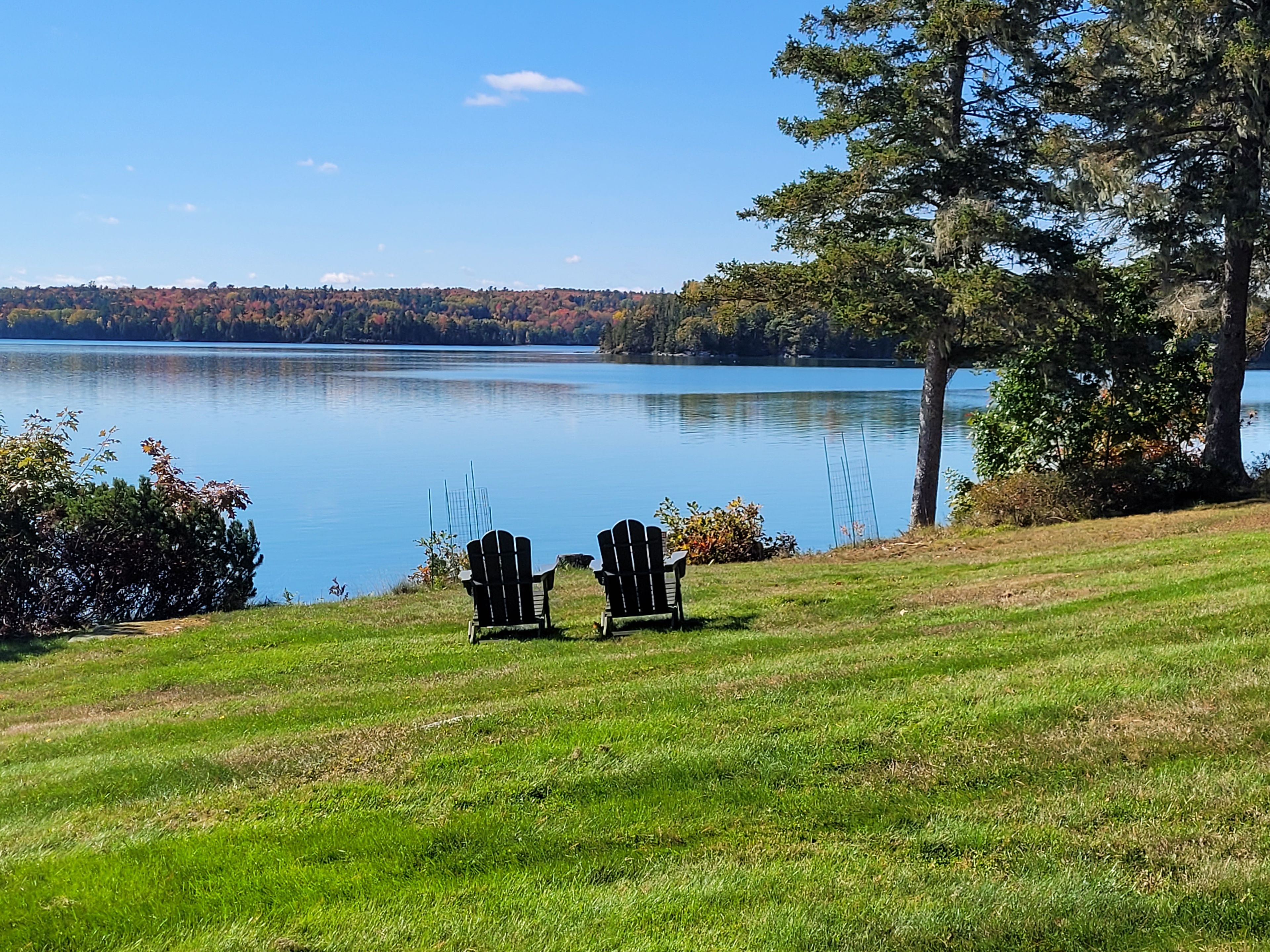 Une zone herbeuse avec deux fauteuils Adirondack donnant sur un lac paisible et une colline boisée à l’horizon.