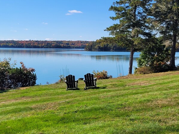 A grassy area with two Adirondack chairs facing a calm lake and a forested hill in the distance.