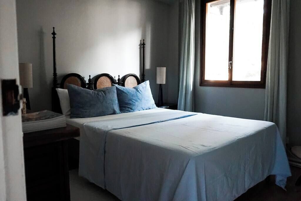 Un dormitorio con cama, mesita de noche, lámpara y ventana con cortinas.