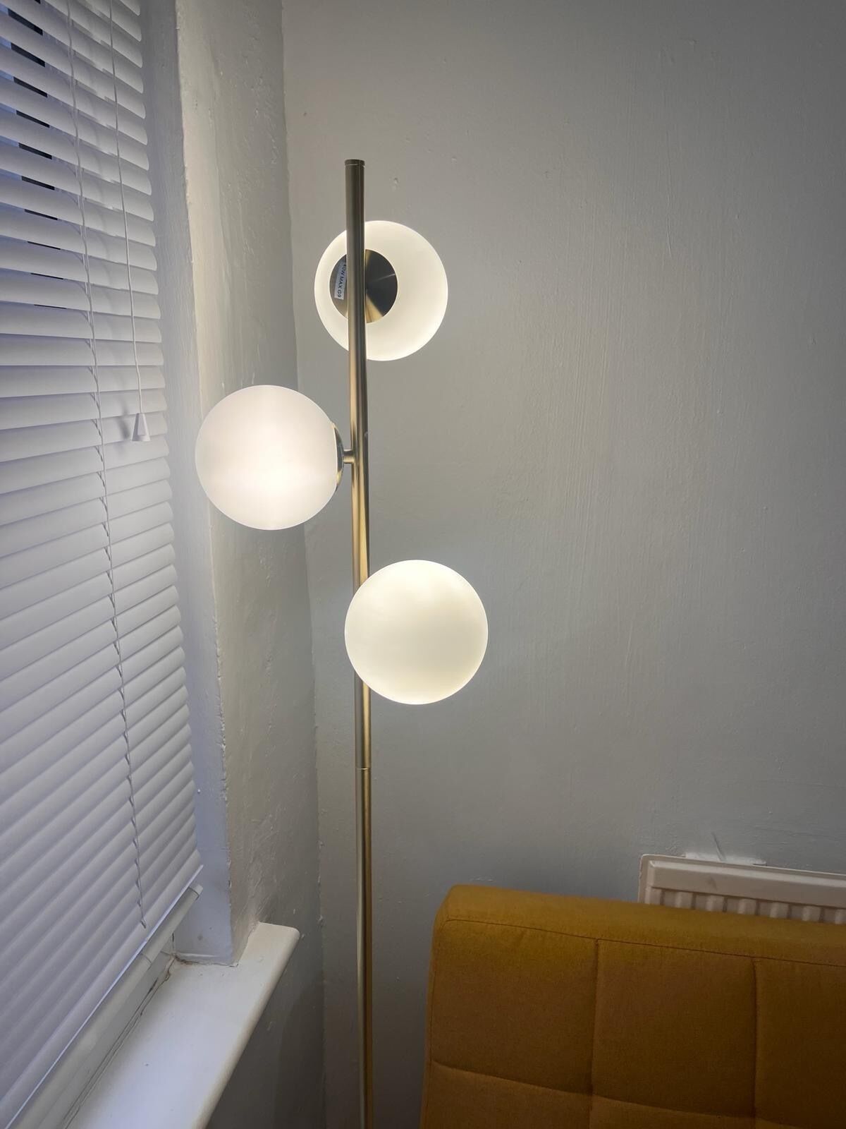 Eine Stehlampe mit drei kugelförmigen Lampenschirmen in einem Zimmer, das eine Heizung und ein Fenster mit Jalousien hat.