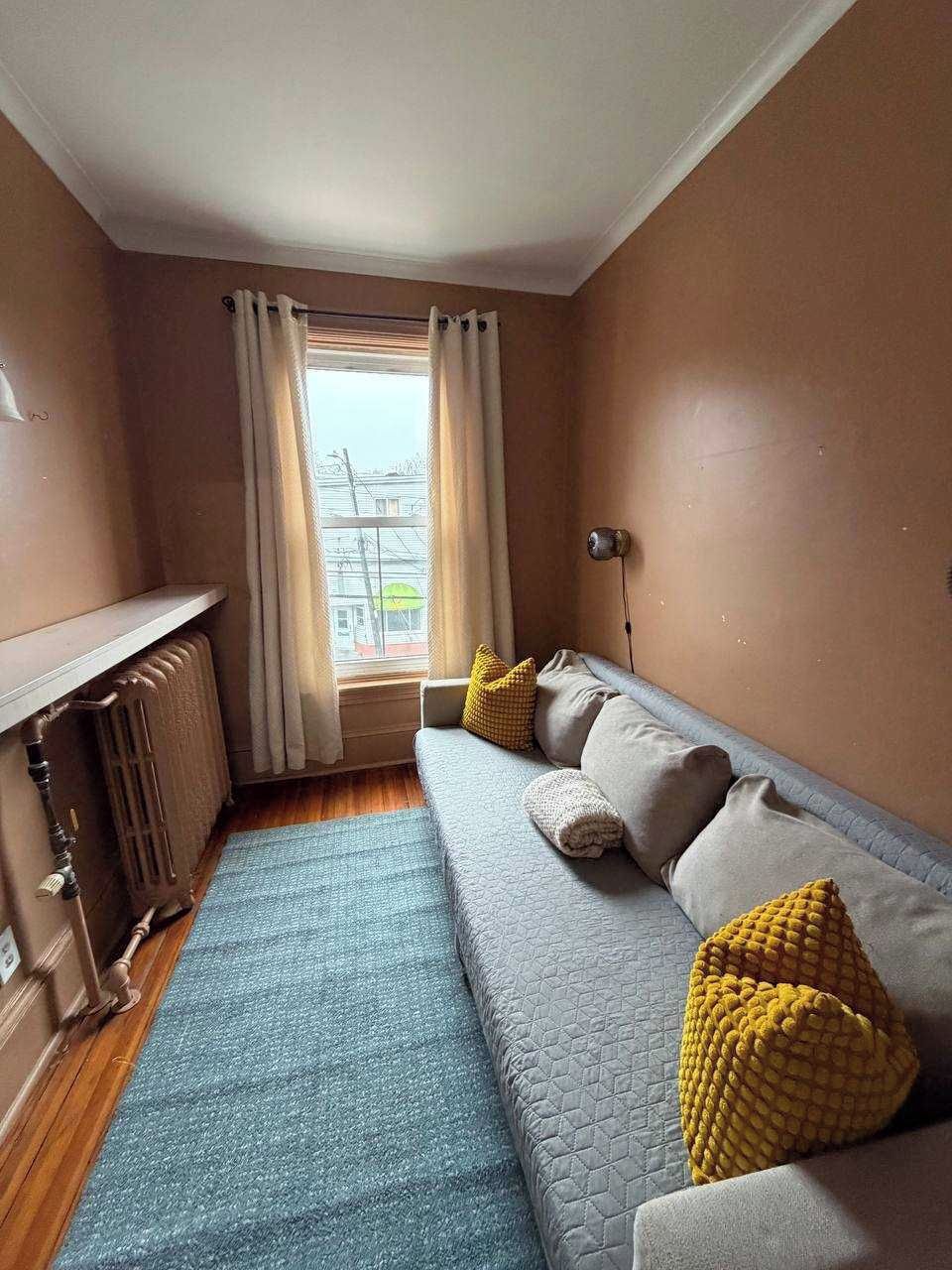 A room with a sofa, a radiator, a window with curtains, and a view of buildings outside.