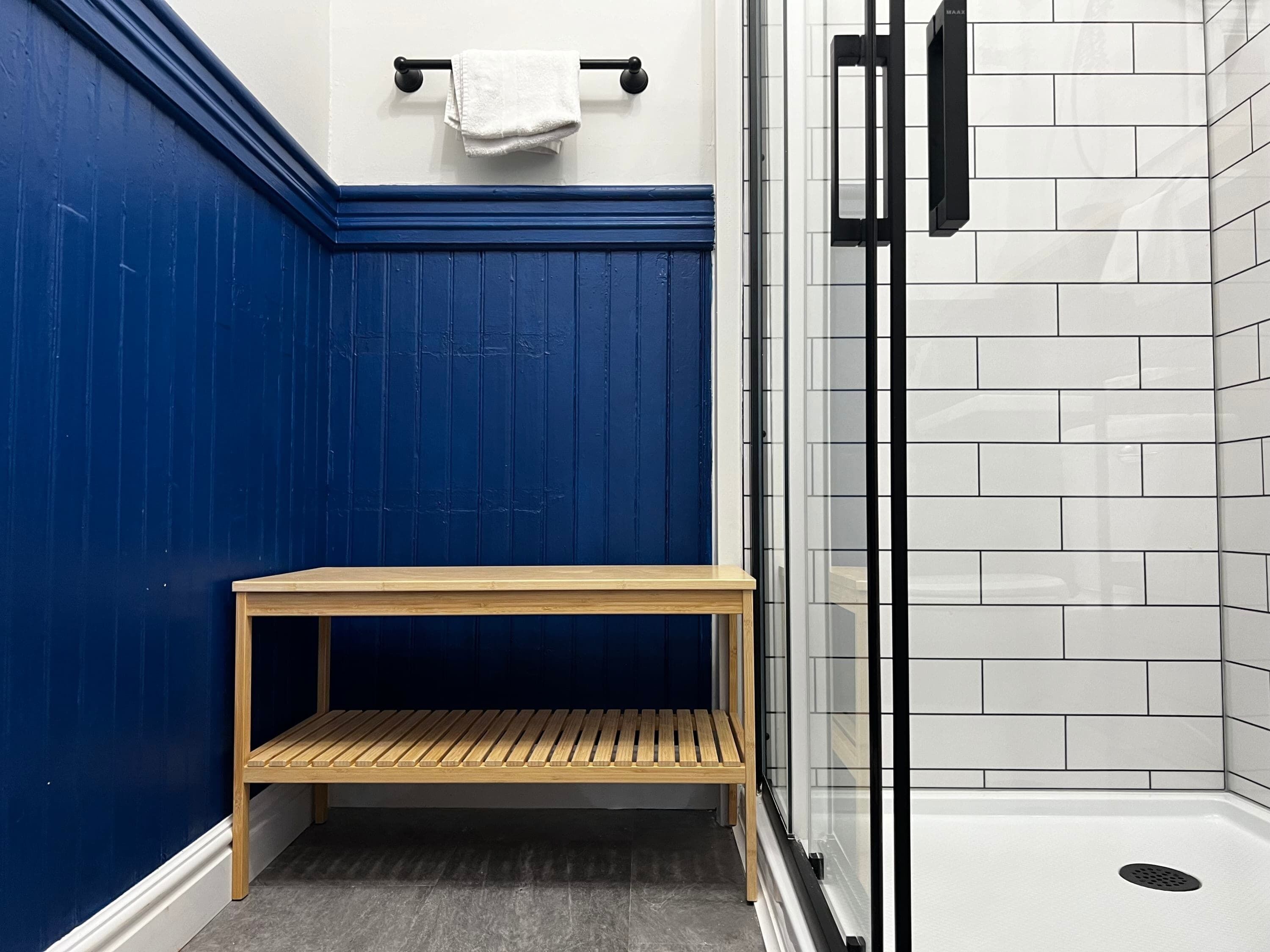 Ein Badezimmer mit blauer Wand, einer Holzbank, einem weißen Handtuch und einer Glasdusche.