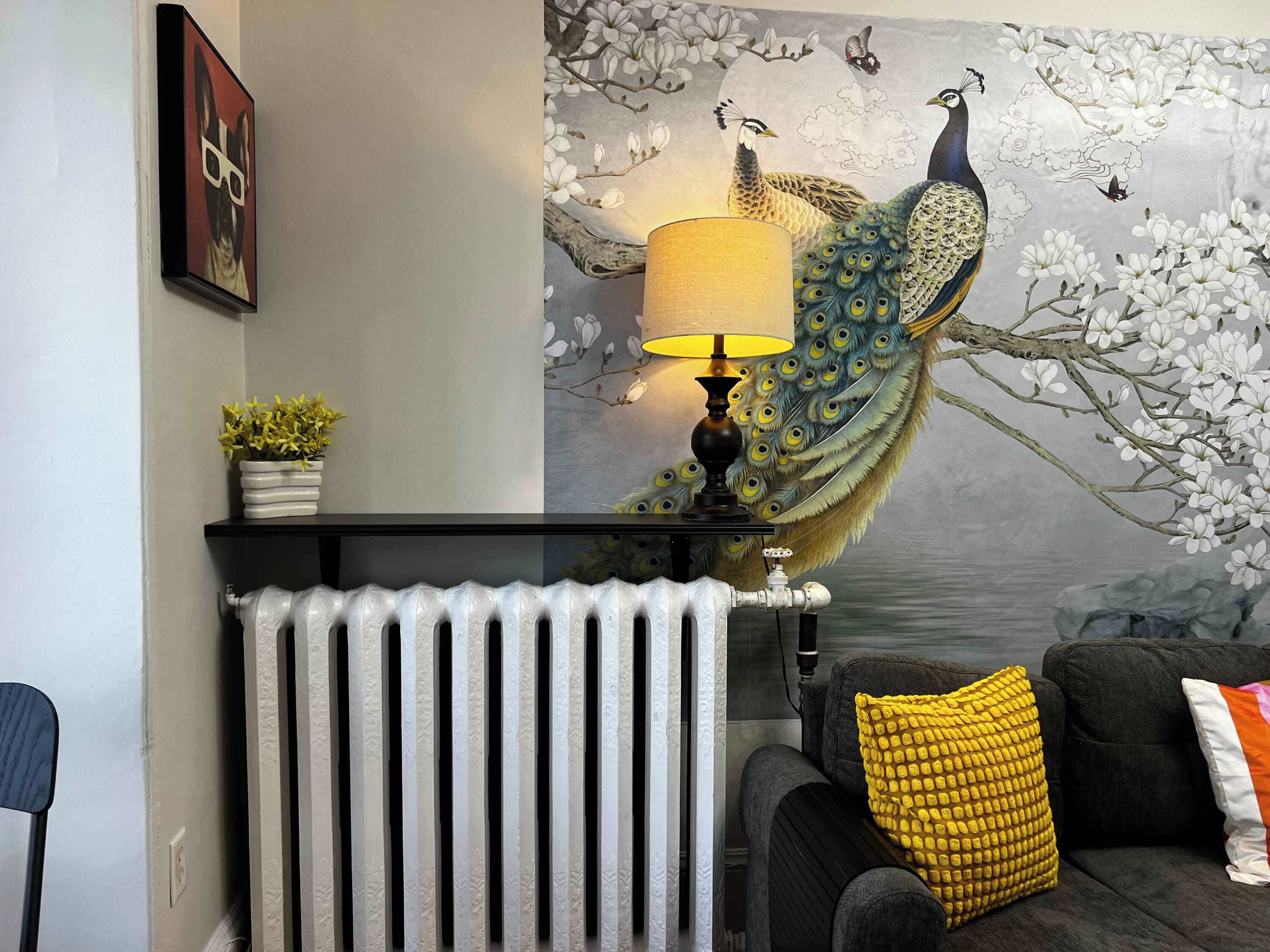 A living room with a radiator, a lamp, a sofa with cushions, a framed artwork, and a small plant on a shelf.
