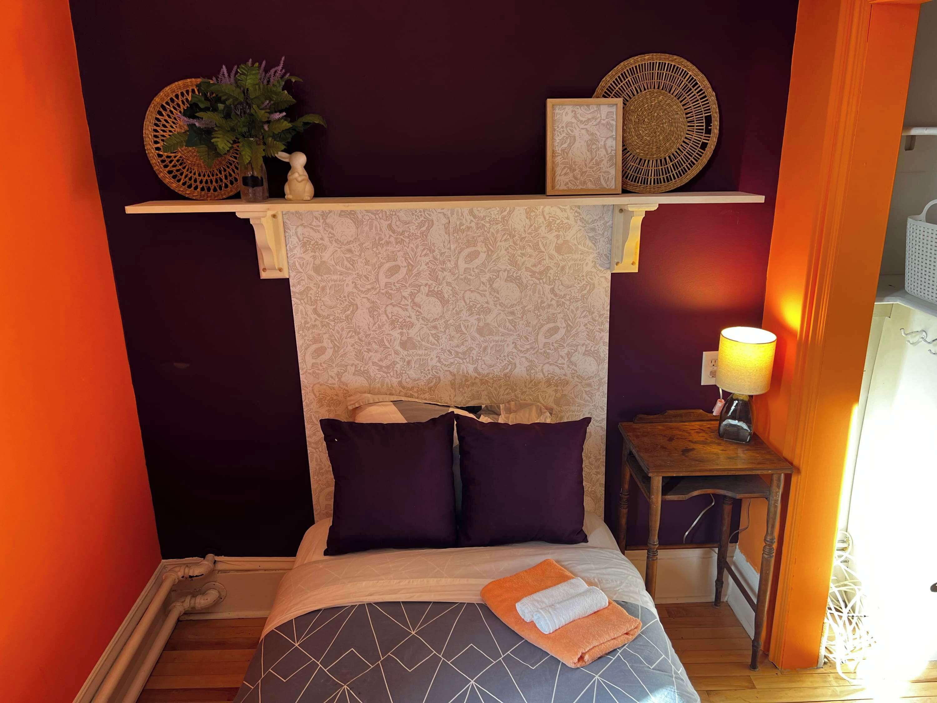 Un dormitorio con cama, almohadas, una mesita de noche con lámpara, estante con adornos y pared con papel tapiz estampado.