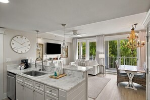 A modern kitchen with a large island, a dining area with a table and chairs, and a balcony with a view of greenery.