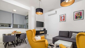 A modern living room with a sofa, yellow armchairs, a dining table with chairs, a TV mounted on the wall, and artwork on the walls.