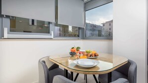A modern dining area with a glass table, grey upholstered chairs, and a breakfast setting with fruits, pastries, and orange juice.
